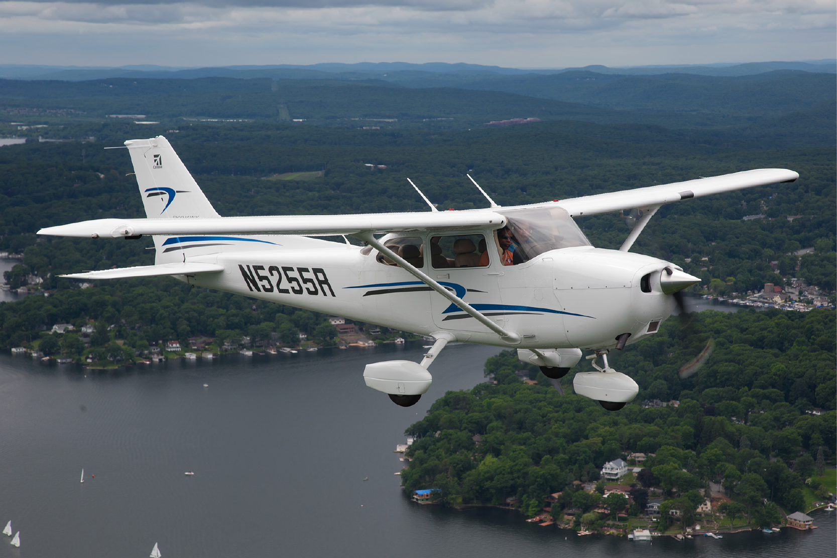 Single-engine white Cessna light airplane (N5255R) with blue stripes flying over a lake and tree-covered shoreline.