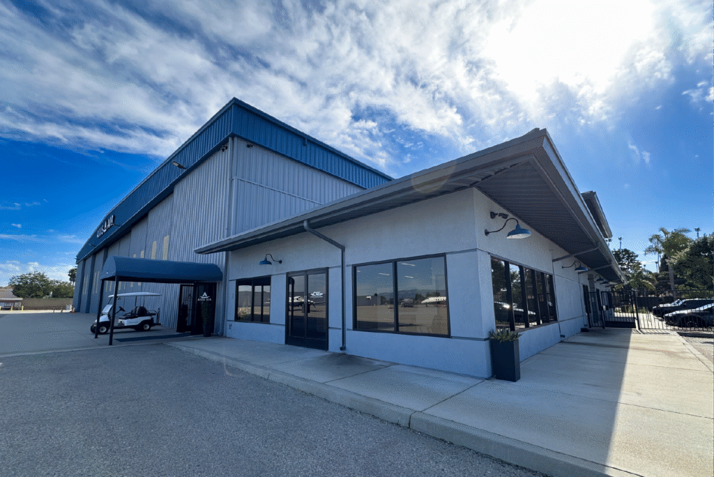 Low industrial building with blue awning and large windows beside a hangar under a bright sky with scattered clouds