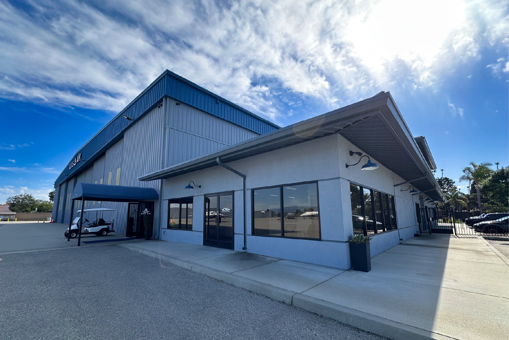 Low industrial building with blue awning and large windows beside a hangar under a bright sky with scattered clouds