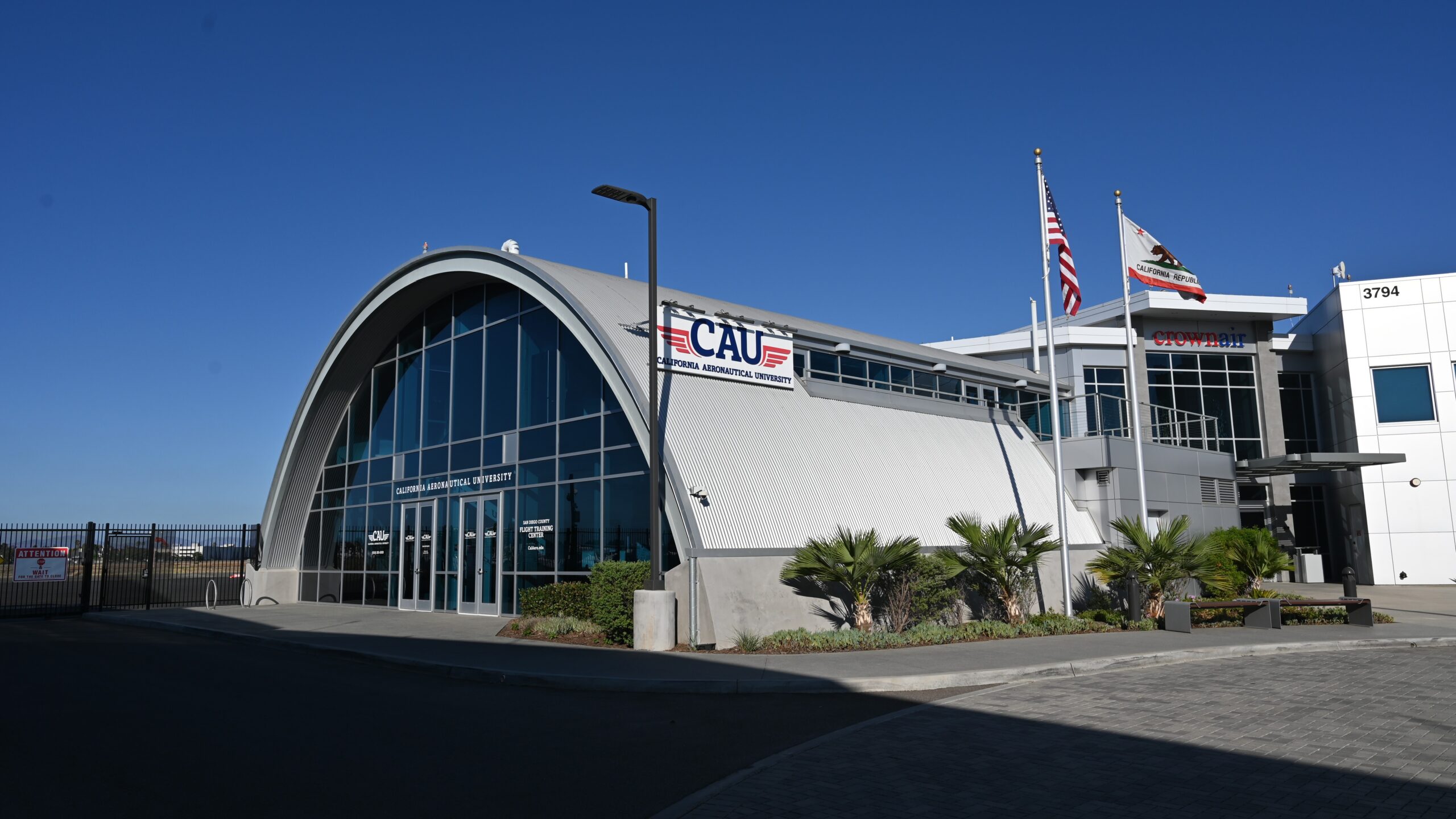 Arched glass-front building labeled CAU (California Aeronautical University) with flagpoles and clear blue sky