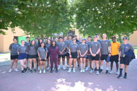 Group of smiling teenage volunteers wearing matching grey shirts standing outdoors under flowering trees.