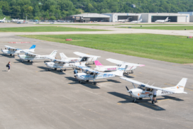 Small single-engine training planes parked on an airport ramp with hangars and grassy taxiways in the background.