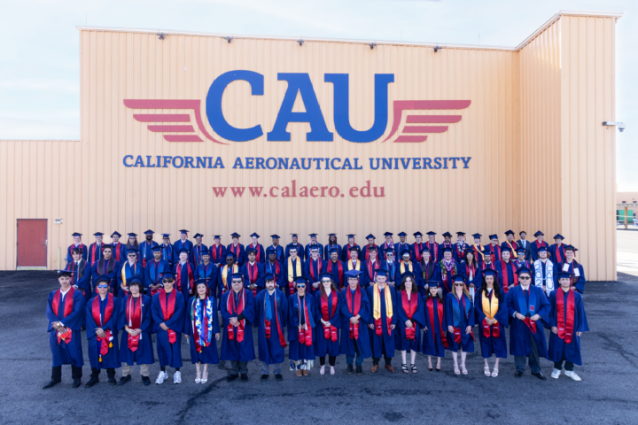 Large group of diverse university graduates in blue caps and gowns with red stoles standing in rows in front of a beige hangar wall painted with a large blue CAU logo and "California Aeronautical University" and the website address.