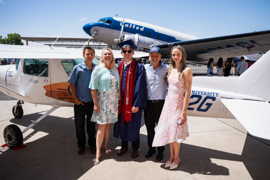 Graduate in cap and gown smiling with family in front of two small airplanes on sunny tarmac