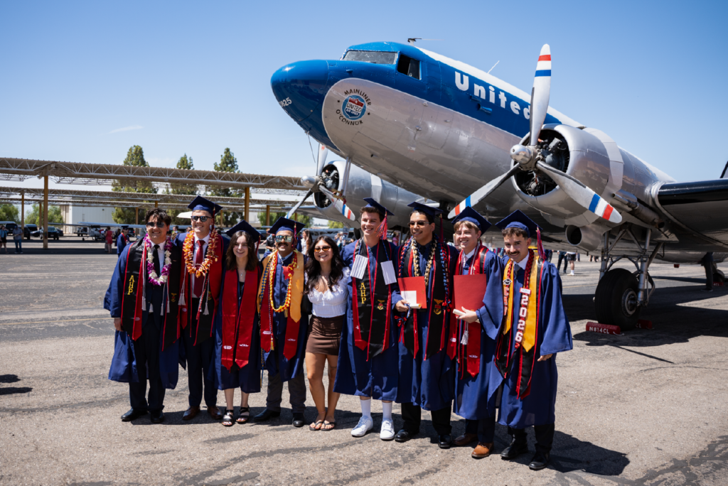 Nine smiling graduates in caps and gowns posing in front of a vintage United Airlines propeller airplane